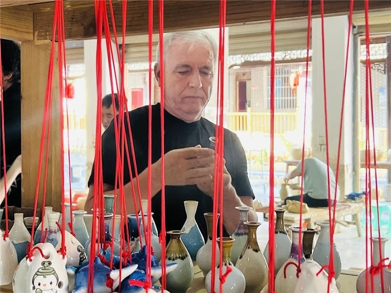 鄉(xiāng)愁如同一根根紅線，將游子與故鄉(xiāng)永遠地牽連到一起?；逄账囄幕瘎?chuàng)意一條街的母氏陶藝館內，紅線吊著陶瓶，思緒游走萬里。摩爾多瓦“信息標簽”通訊社社長亞歷山大·塔納斯端詳著館內展示的陶藝作品，并挑選出心儀的小擺件帶回摩爾多瓦，也帶走花茂村的“鄉(xiāng)愁”。（張亞欣攝）